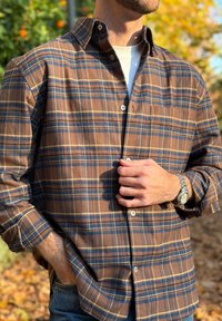 Homme portant une chemise à carreaux marron et bleue sur un t-shirt blanc, une main dans la poche, l'autre ajustant un bouton de la chemise, à l'extérieur avec un feuillage d'automne.