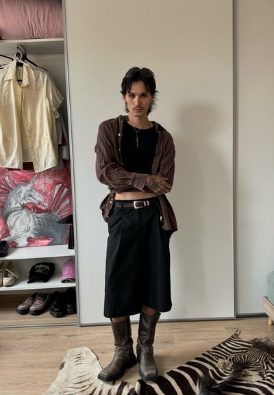 Una persona vestida con una camisa de botones marrón sobre una blusa negra, una falda negra y botas marrones, de pie sobre una alfombra con diseño de cebra.