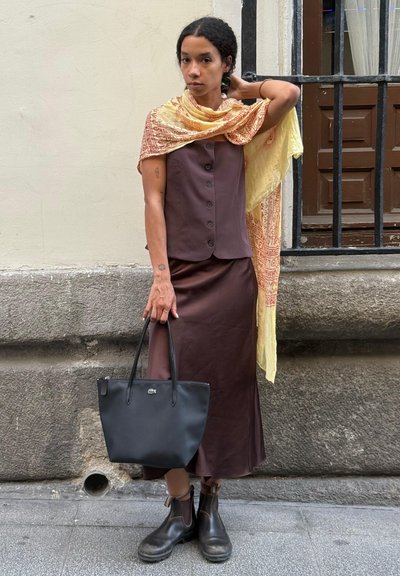 Conjunto marrón de dos piezas que incluye una blusa sin mangas con botones, una falda larga y una bufanda amarilla con estampado. Bolso de piel negra y botas.