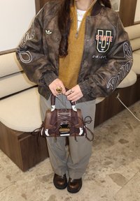 Άτομο που φοράει καφέ αθλητικό μπουφάν, μουσταρδί πουλόβερ, γκρι παντελόνια και καφέ παπούτσια, κρατώντας μια καφέ τσάντα με μοτίβα και λεπτομέρειες με κορδόνια.