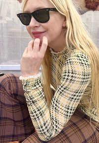 Donna bionda con occhiali da sole neri, indossa una maglia giallo-verde a motivo pied-de-poule e pantaloni marroni a quadri, appoggia il mento sulla mano e sorride.