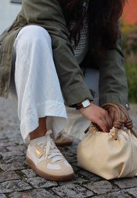 Baskets beige avec panneaux texturés et accents blancs, associées à un sac tressé camel. La personne est accroupie sur un pavé en pierre.