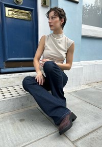 Μπεζ αμάνικο μπλουζάκι, σκούρο μπλε φαρδύ τζιν παντελόνι, σκούρες καφέ πλατφόρμες. Καθισμένη σε σκαλιά με μια μπλε πόρτα στο παρασκήνιο.