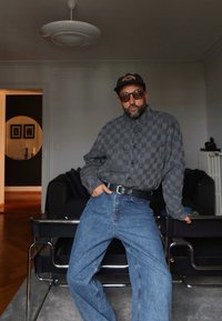 Mann med solbriller, caps, rutete skjorte og blå jeans lener seg mot en stol i stuen med mørk sofa og rundt veggspeil i bakgrunnen.
