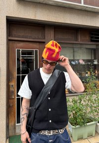 Czerwona wełniana czapka z żółtymi płomieniami, czarna dzianinowa kamizelka, biała koszulka, czarna skórzana torebka na ramię oraz niebieskie dżinsy z paskiem w panterkę.