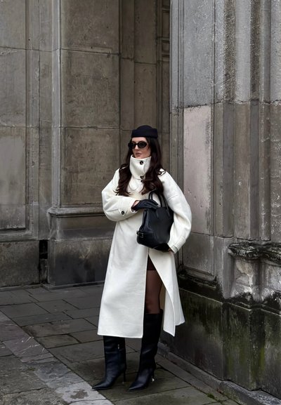 Abrigo blanco de gran tamaño con cuello alto, guantes negros y bolso estructurado negro. Botas negras hasta la rodilla y gafas de sol oscuras complementan el look.