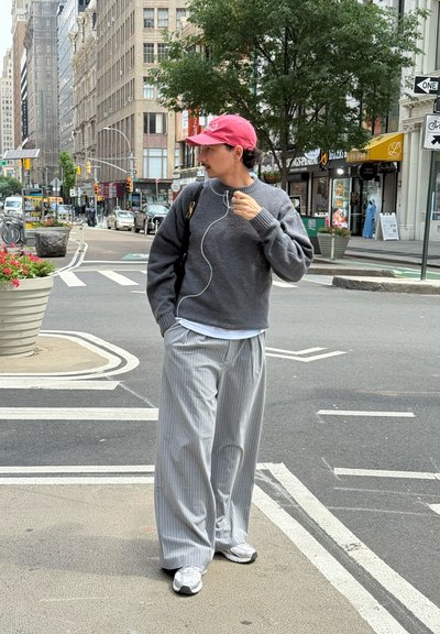 Suéter gris, pantalones anchos a rayas grises, camiseta blanca debajo. Gorro rosa, auriculares y zapatillas blancas. Escena urbana.
