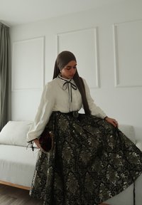 Femme portant un hijab marron foncé, un chemisier blanc avec un nœud, et une jupe ornée de motifs floraux noirs et dorés, debout dans une pièce minimaliste avec un canapé.