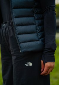 Osoba v čiernej vypchanej veste a dlhým rukávom s otvorom na palce, v čiernych nohaviciach s logom "The North Face", stojí vonku.