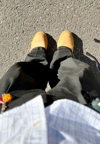 Brūni ādas zābaki uz melnajām biksēm, ar daļēji redzamu rūtainu baltu krekli. Virsma ir teksturēts asfalts.