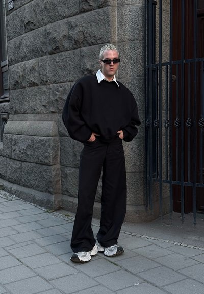 Sudadera negra de gran tamaño con cuello blanco, pantalones anchos negros y zapatillas plateadas, contrastando con una pared de piedra gris texturizada.