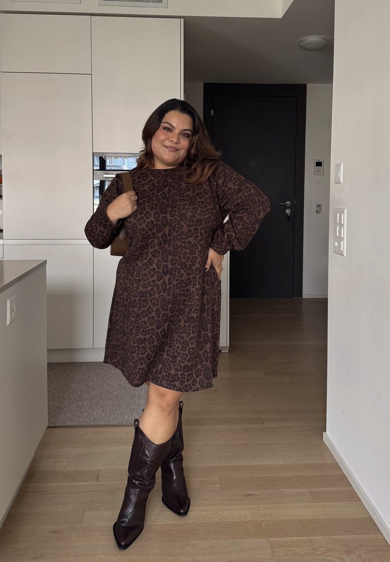 Robe à imprimé léopard marron avec des manches longues, coupe décontractée, associée à des bottes en cuir marron à bout pointu. Fond intérieur moderne.