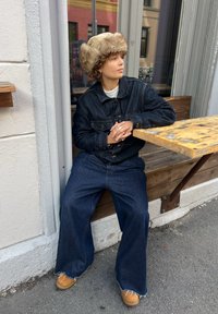 Jeansowa kurtka z przednimi kieszeniami, luźne dżinsowe spodnie i beżowy kapelusz z futrzanym wykończeniem. W tle drewniana ławka i chodnik.