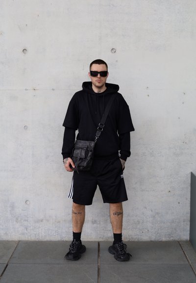 Sudadera negra de gran tamaño con mangas cortas, pantalones cortos negros con rayas blancas y zapatillas deportivas negras gruesas. Lleva gafas de sol y un bolso texturizado.