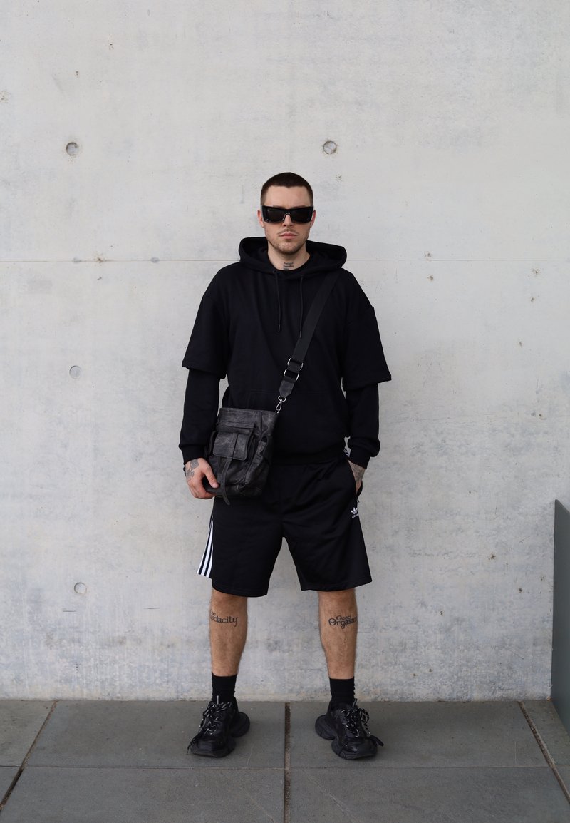 Μαύρη oversized που hoodie με κοντά μανίκια, μαύρα σορτς με άσπρες ρίγες και χοντρές μαύρες αθλητικές σκαρπίνες. Φοράει γυαλιά ηλίου και μια υφασμάτινη τσάντα.