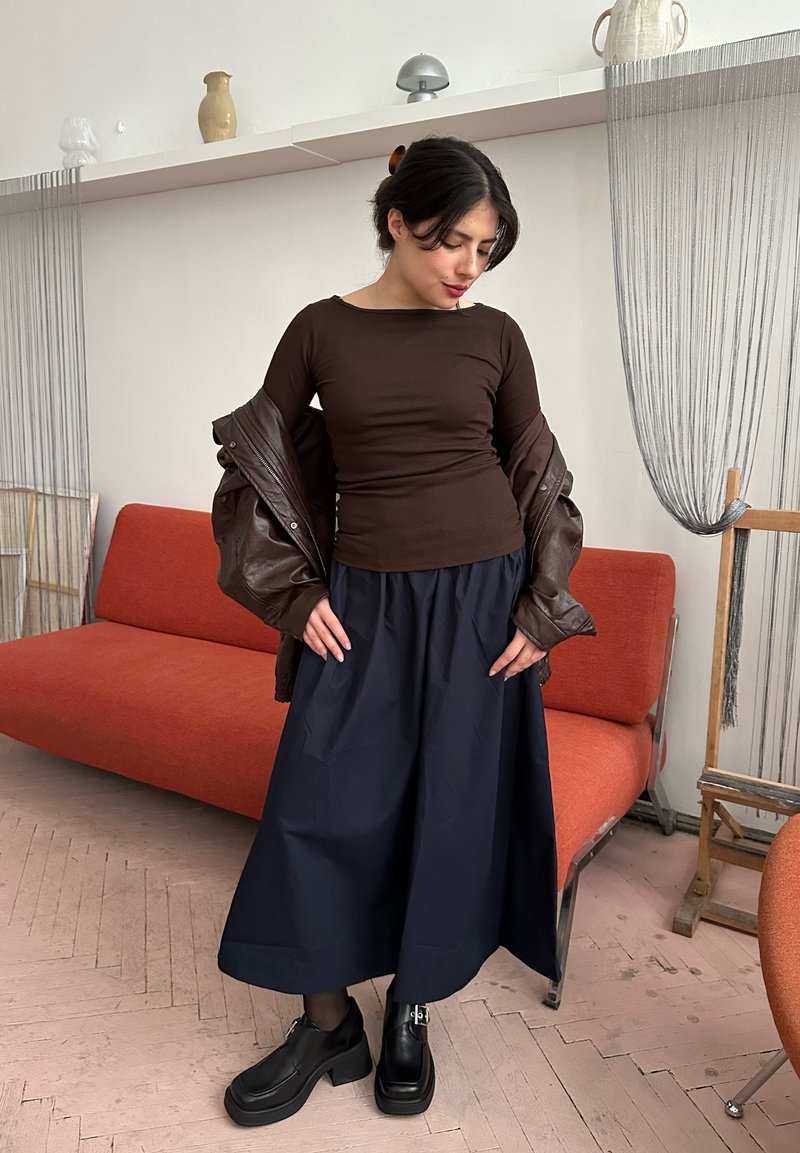 Femme ajustant une veste marron foncé sur un haut marron moulant, portant une jupe large bleu marine et des chaussures plateformes noires, debout près d'un canapé orange à l'intérieur.