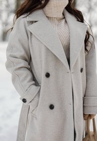 Femme portant un manteau en laine beige et un pull à col roulé, tenant un sac à main en cuir camel en extérieur par temps neigeux.