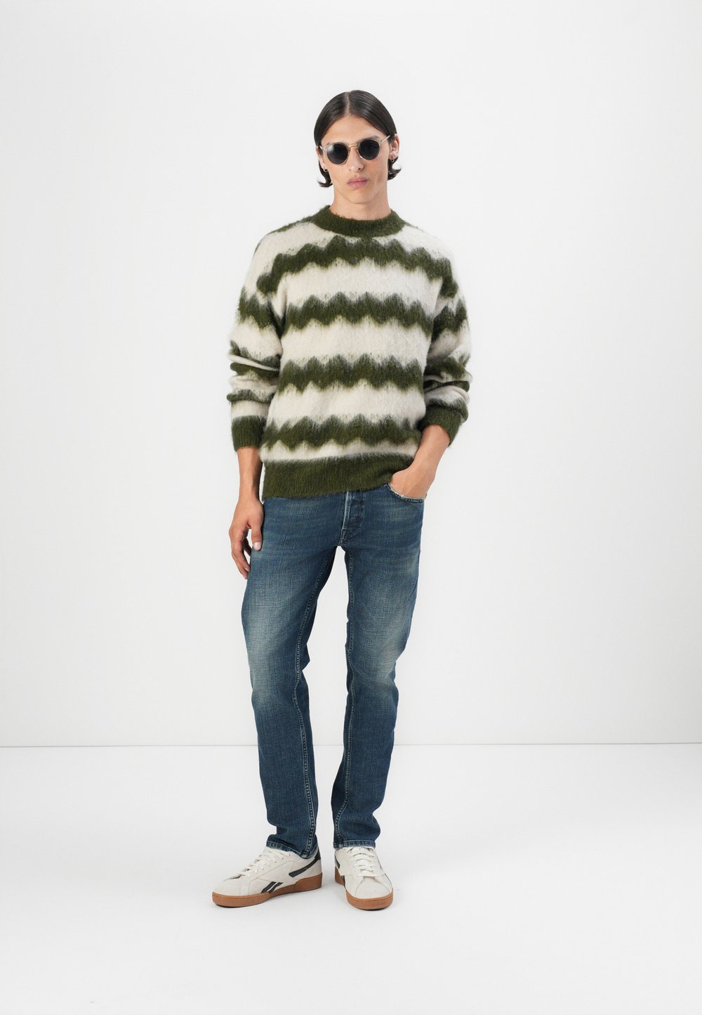 Kreminės vilnos mišinio megztinis su žaliu zigzago raštu, apvali iškirptė ir elastingos rankovės. Suderintas su mėlynomis džinsinėmis kelnėmis ir kreminėmis sportbačiais.