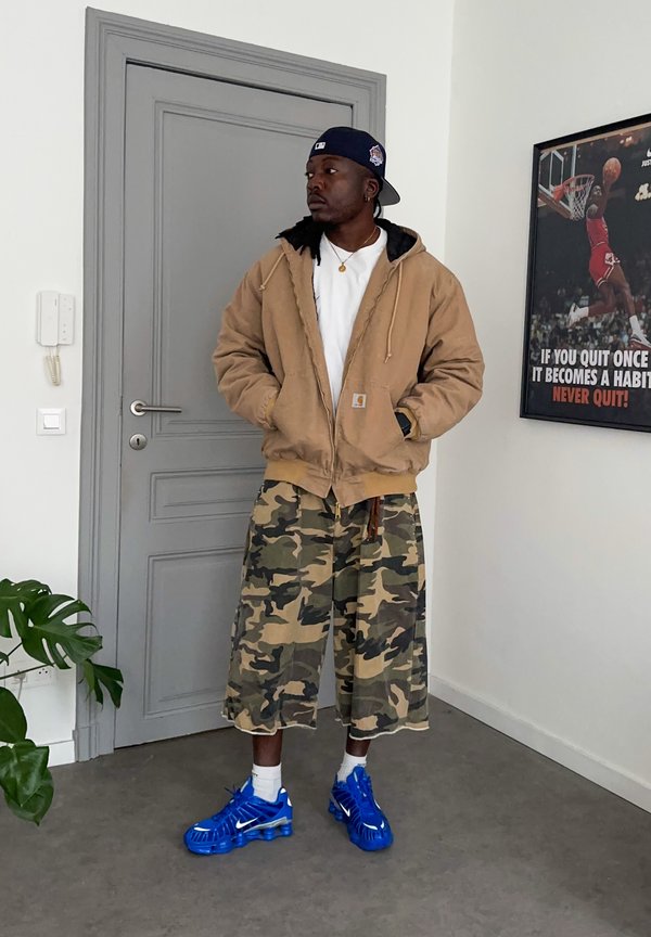 Hombre con chaqueta marrón claro, pantalones cortos de camuflaje, zapatillas azules y gorra azul marino al revés, está de pie en el interior cerca de una puerta gris y un póster motivacional de baloncesto.