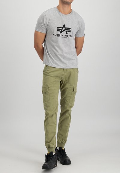 Camiseta de algodón gris con gráfico de "ALPHA INDUSTRY", combinada con pantalones cargo verdes con bolsillos laterales y puños elásticos, usados con zapatillas negras.