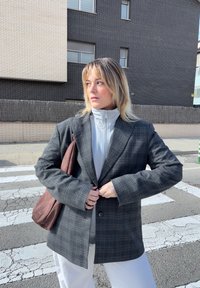 Blazer a quadre grigi con maniche larghe, chiusura con un solo bottone, indossato sopra un top zip grigio chiaro; porta una borsa a spalla marrone.