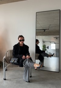 Femme portant un manteau noir, un pantalon gris et des talons, assise jambes croisées sur une chaise rembourrée à côté d'un grand miroir sur pied dans une pièce minimaliste.