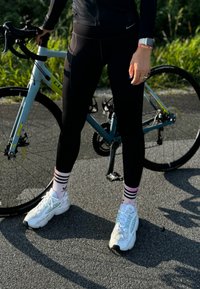 Zwarte sportleggings, witte sneakers met mintaccenten en roze gestreepte sokken, gecombineerd met een blauwe racefiets in een natuurlijke omgeving.
