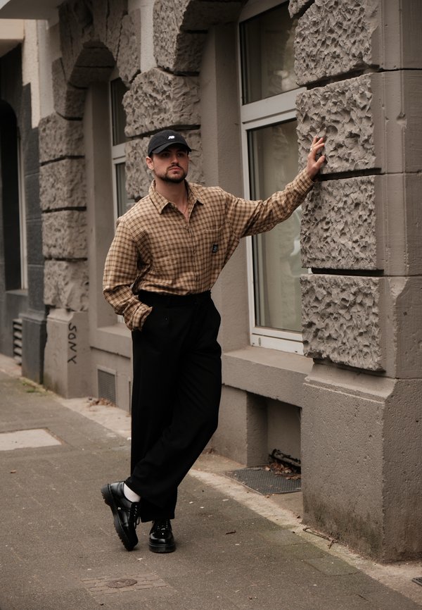 Kockás barna, oversized ing, fekete, bő szárú nadrág és fekete cipő. A férfi egy textúrált kőfal mellett áll, az ablaknál.