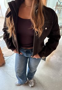 Femme portant une veste noire sur un haut sombre, jean bleu, baskets blanches, debout à l'intérieur avec les mains près de la taille.