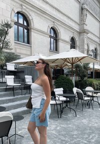 Balta ribota bezplecu blūze, zili džinsu šorti, stāv uz kāpnēm ar ārtelpu sēdvietām un lietussargiem, zaļumi fonā, pilsētas vide.