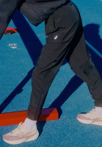 Crne sportske hlače s patentnim zatvaračima na gležnjevima, nošene sa svijetlim cipelama i bijelim čarapama, na plavoj površini sa sjenama.