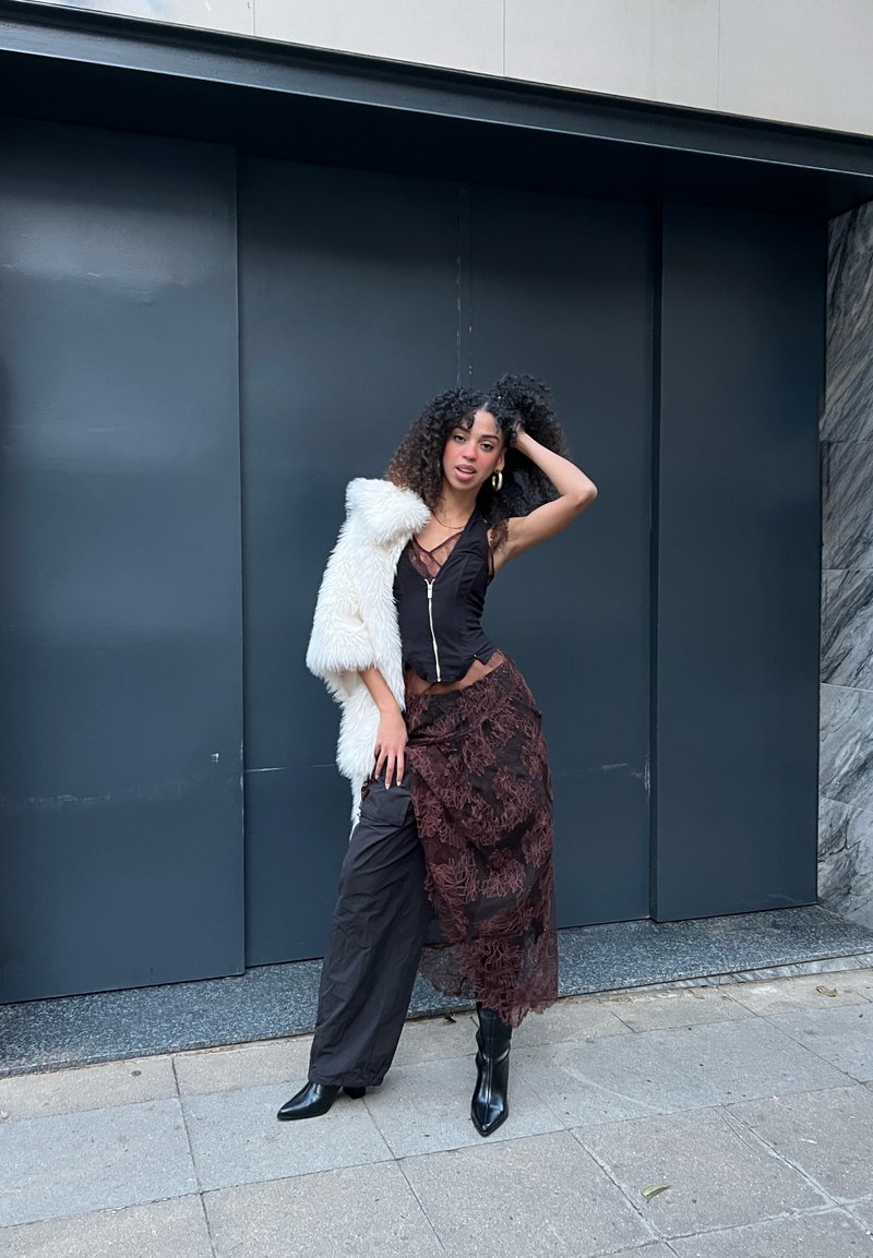 Top nero aderente con zip, gonna marrone a strati con dettagli in pizzo e cappotto di pelliccia bianca. Stivali neri con tacco completano l'outfit.