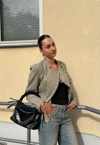 Mujer con chaqueta beige, top negro y vaqueros apoyada en una barandilla metálica, sosteniendo un bolso negro, de pie frente a una pared amarilla texturizada.