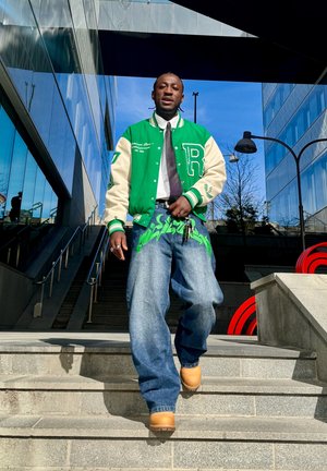 Hombre bajando las escaleras con una chaqueta universitaria verde y blanca, vaqueros holgados con diseño verde, corbata y botas marrones en un entorno urbano.