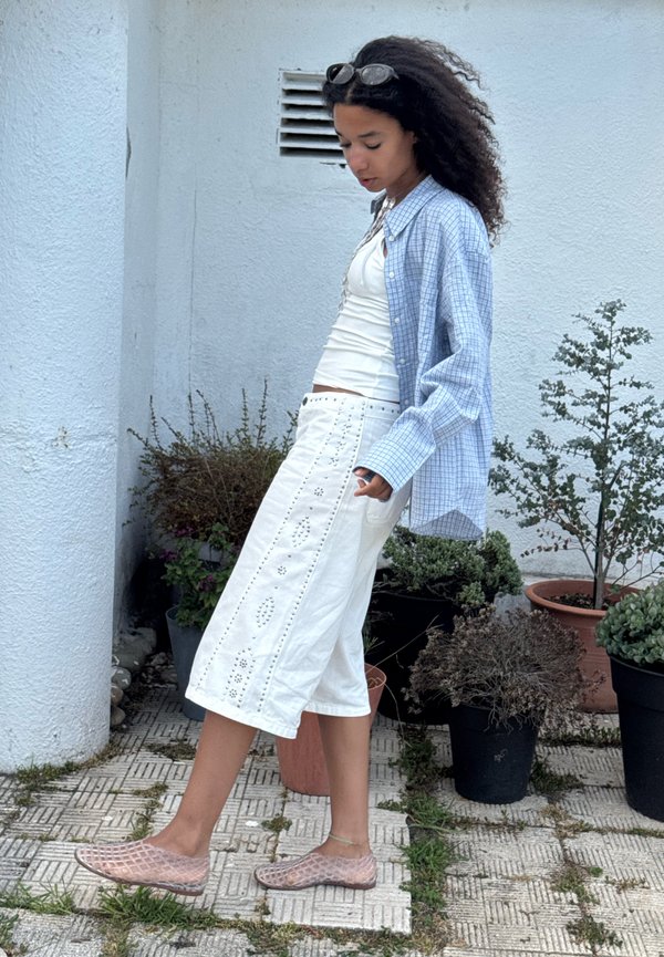 Mujer con cabello rizado que lleva gafas de sol en la cabeza, camiseta de tirantes blanca, falda blanca bordada, camisa azul a cuadros y zapatos de malla rosa camina al aire libre cerca de plantas en macetas.
