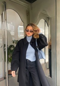 Casaco oversized preto, suéter de gola alta azul claro, jeans pretos, pequena bolsa azul, óculos de sol; superfícies refletivas e suaves num ambiente moderno.