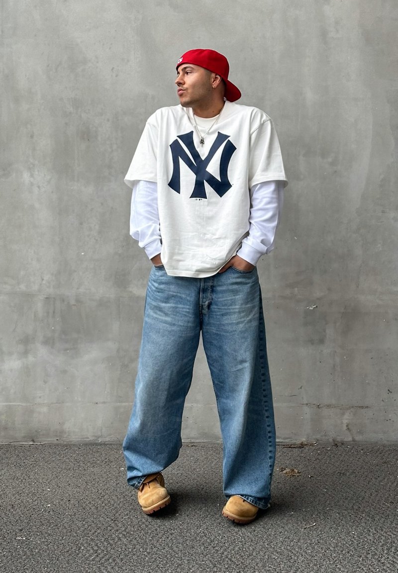Λευκό T-shirt με σκούρο μπλε λογότυπο "NY", στρωμένο πάνω από λευκό πουκάμισο μακρύ μανίκι. Ανοιχτό μπλε φαρδύ τζιν και καφέ παπούτσια, με κόκκινο καπέλο.