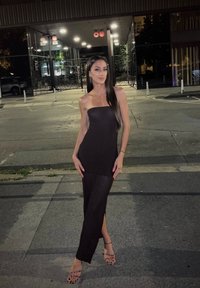 Rochie maxi neagră cu textură rigată, cu design fără bretele și fanta laterală. Poartă sandale cu toc transparente, amplasată pe un fundal urban de noapte.