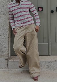 Polo blanc à manches longues avec des rayures rouges, associé à un pantalon large beige et des chaussures marron, se tenant sur une surface en pierre près d'une porte grise.