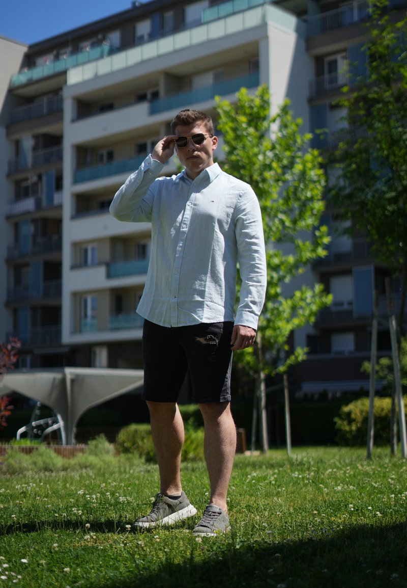 Mann trägt weisses langärmeliges Hemd, schwarze Shorts und Sonnenbrille, steht auf Gras mit Bäumen und einem Wohngebäude im Hintergrund.
