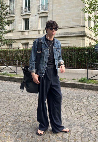 Joven con gafas de sol, chaqueta vaquera, pantalones negros anchos, chanclas, llevando una bolsa negra, caminando por una calle adoquinada cerca de un edificio beige.