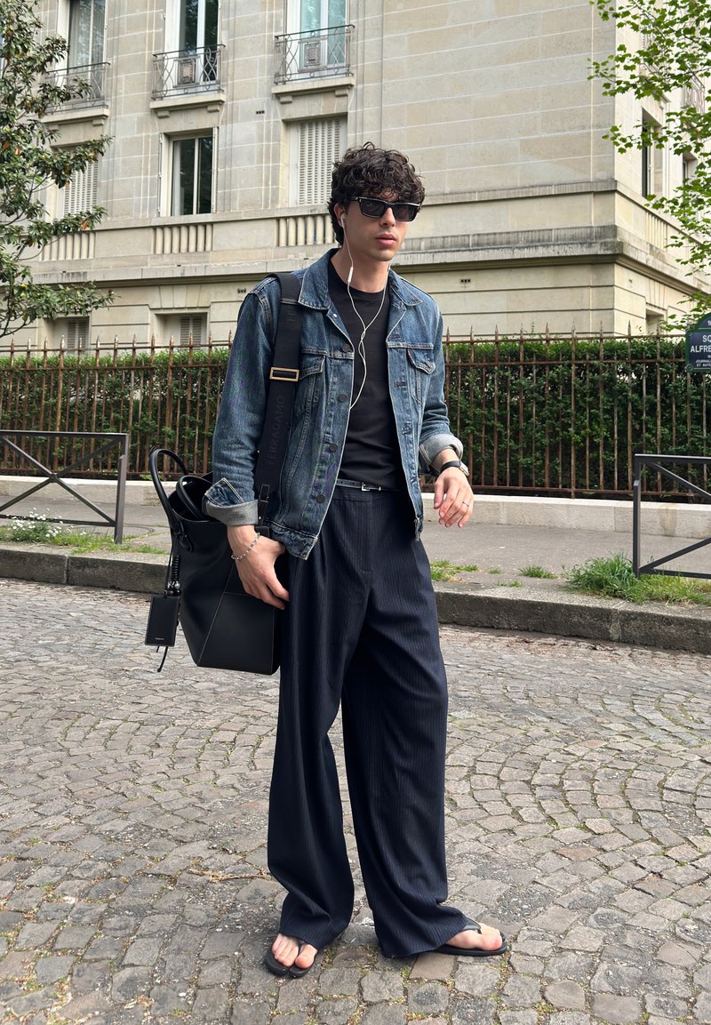 Jeune homme portant des lunettes de soleil, une veste en jean, un pantalon large noir, des tongs, portant un sac noir, marchant sur une rue pavée près d’un bâtiment beige.
