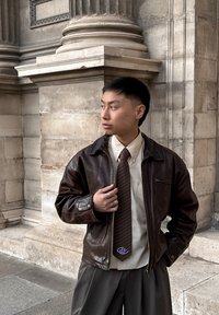 Bruine leren jas over een licht shirt en gestreepte stropdas, met een zak en decoratieve ring. Grijze broek; architectuur op de achtergrond.