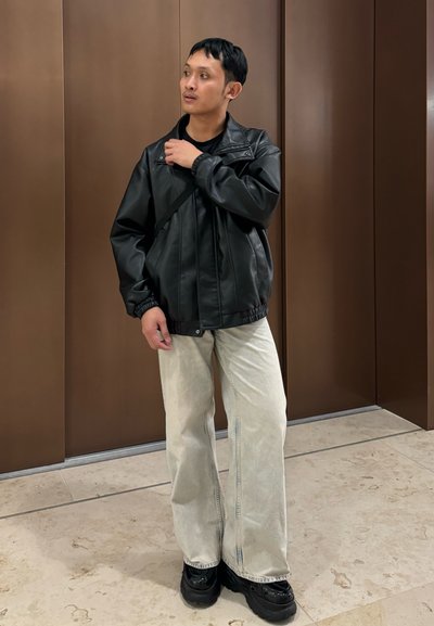 Chaqueta de cuero sintético negra con cuello alzado, llevada sobre una blusa negra, combinada con unos pantalones de mezclilla de pierna ancha beige claro y zapatos negros gruesos.