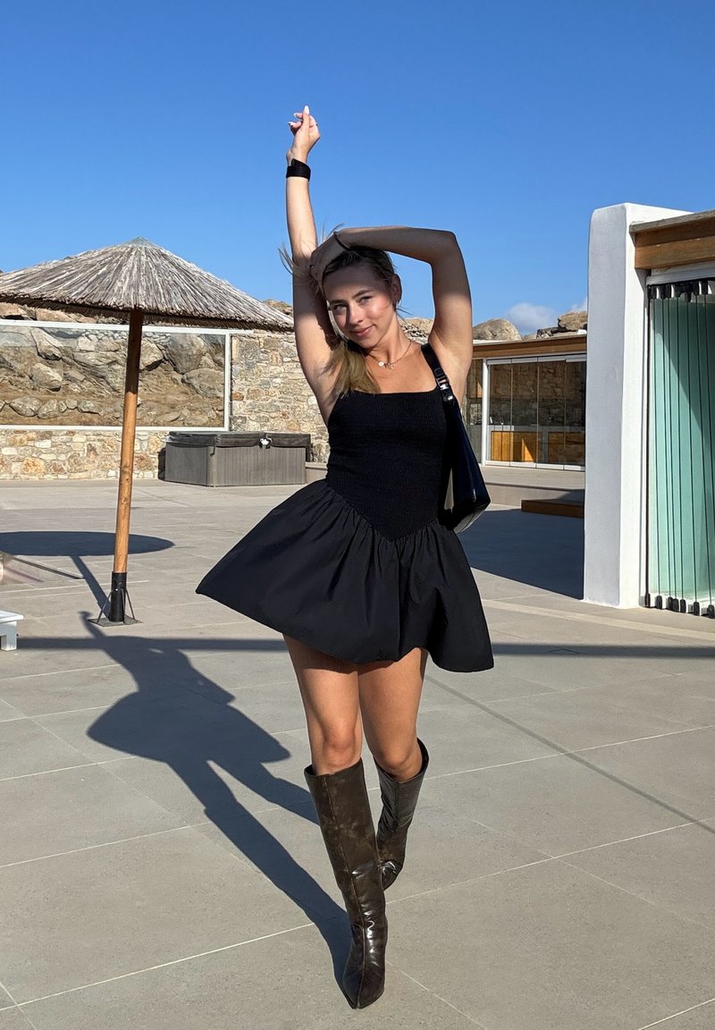 Robe noire ligne A avec tissu texturé, corsage ajusté et jupe évasée. Bottes longues marron et sac à épaule complètent le look. Environnement extérieur.