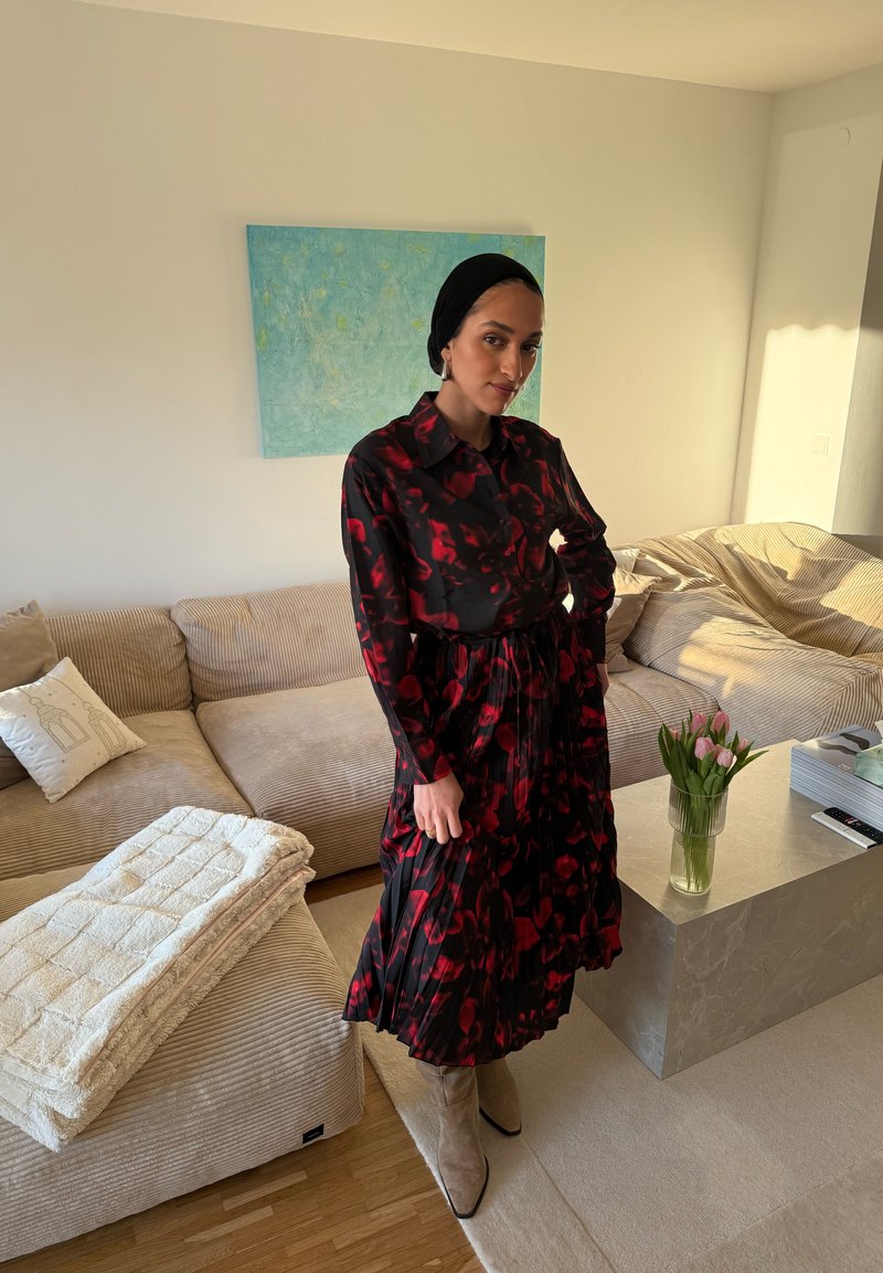 Femme en robe noire à fleurs rouges et bottes beige se tenant dans un salon beige près d'un canapé et d'une table basse en marbre avec des tulipes roses.
