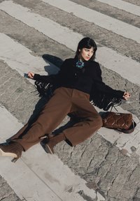 Femme allongée sur un passage pour piétons pavé, portant un pantalon marron, des bottes à talons marron, une veste noire à franges et une grande broche bleue, avec un sac marron à proximité.