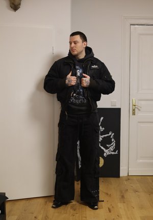 Hombre con tatuajes vistiendo pantalones cargo negros, sudadera con capucha gráfica y chaqueta negra, de pie en el interior cerca de puertas blancas y arte de esqueleto.