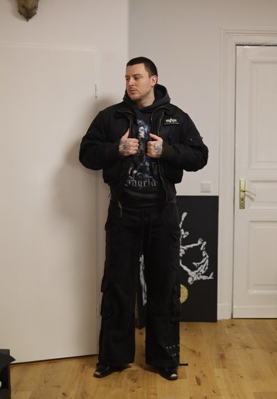 Hombre con tatuajes vistiendo pantalones cargo negros, sudadera con capucha gráfica y chaqueta negra, de pie en el interior cerca de puertas blancas y arte de esqueleto.