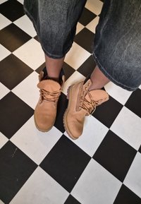 Brune arbeidssko med semelskinn, snøring og gummisåler. Båret med avkuttede, slitte denimbukser på et rutete gulv.